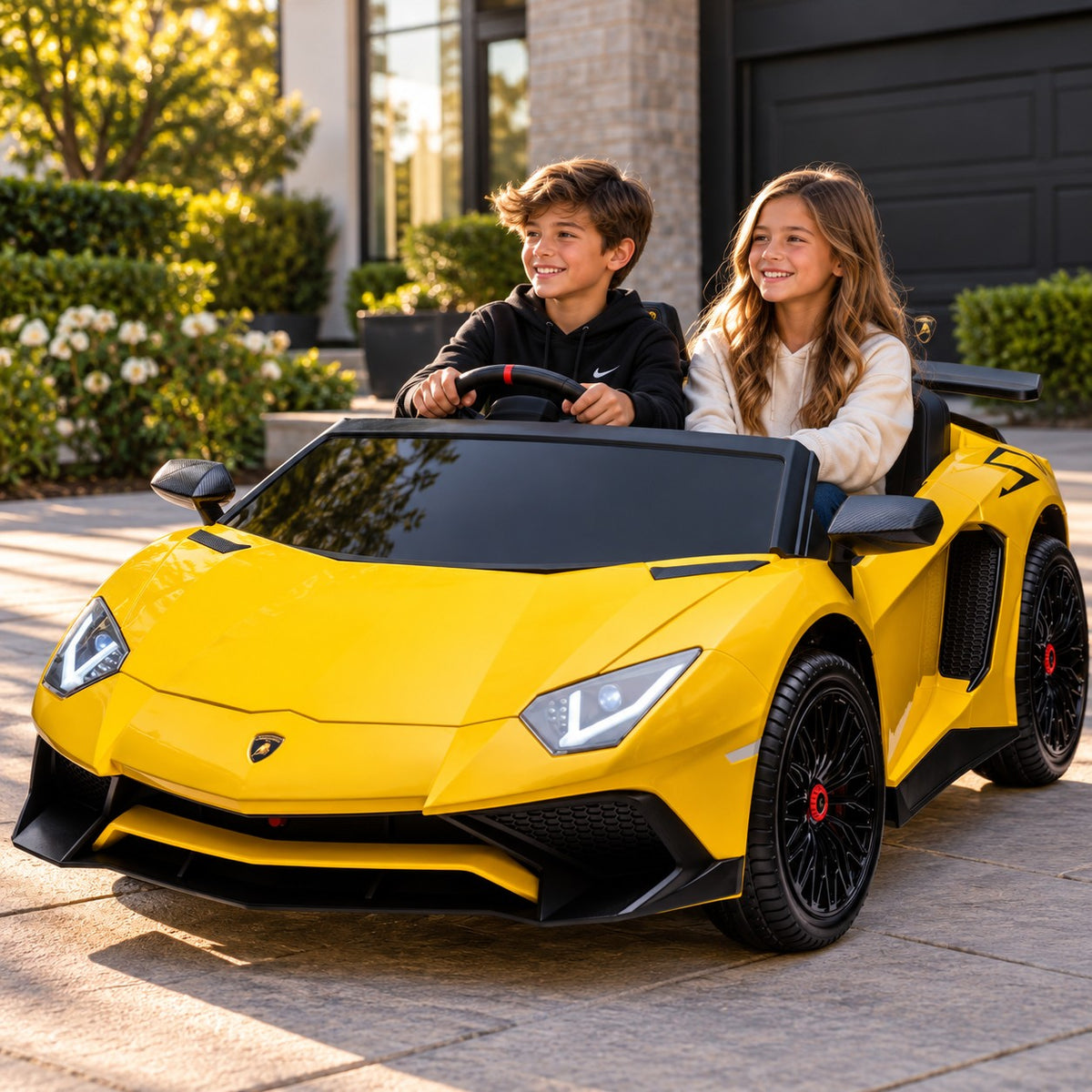Lifestyle Photo of Two Kids Riding the Yellow Lamborghini Aventador XXL 24V Big Kids Ride-On Car on a Home Driveway