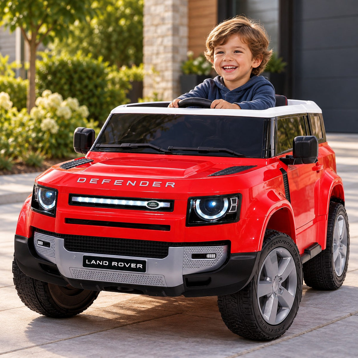 Lifestyle Photo of a Child Driving the Red Land Rover Defender 12V Kids Ride-On Car with LED Lights on a Home Driveway